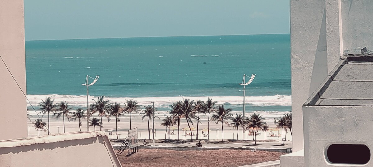The image showcases a view of the beach featuring calm turquoise waters and a sandy shoreline lined with palm trees. Sunbathers can be seen enjoying the beach in the distance, while a clear blue sky completes the serene coastal scene.