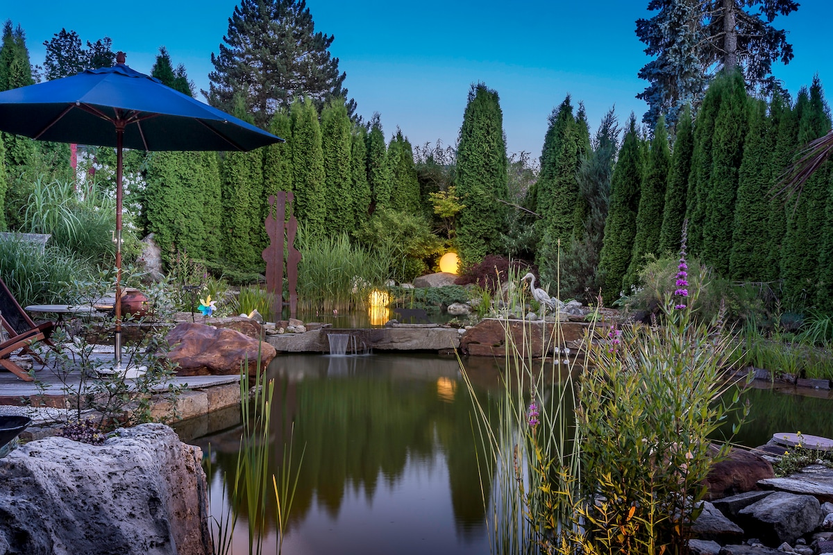 A serene garden pond is framed by lush greenery and towering shrubs. Soft lighting is visible in the background, creating a calming ambiance. An umbrella shades a seating area nearby, while ornamental plants enhance the natural beauty surrounding the water.