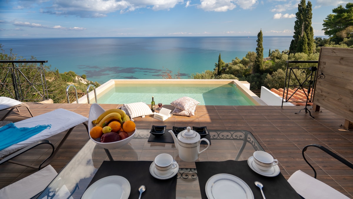 A dining table is set with white dishes and utensils, positioned near a spacious infinity pool overlooking the sea. Comfortable loungers are arranged nearby, accompanied by a selection of fresh fruits. Lush greenery and distant trees create a serene backdrop against a clear blue sky.