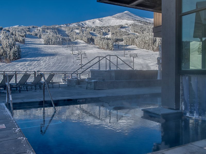 An outdoor hot tub is surrounded by frost-covered trees, with stunning views of snow-covered ski slopes in the background. Steam rises from the water, creating a serene atmosphere on a crisp winter day. Lounge chairs are positioned nearby for relaxation.