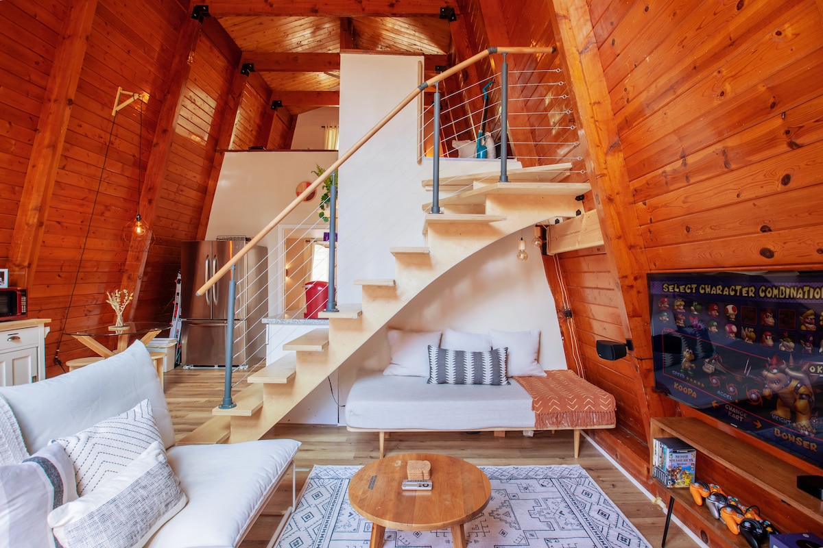 A modern A-Frame interior features high vaulted wood ceilings and a warm wooden wall. A light-colored sofa faces a television and is accompanied by a round coffee table on a patterned rug. A staircase leads to the loft area, adding a spacious element to the living space.