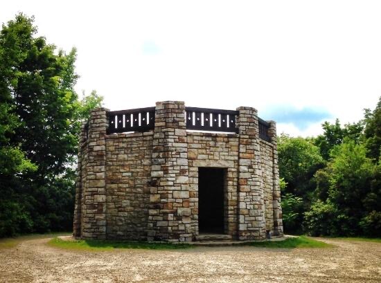 A circular stone structure is surrounded by greenery, featuring a sturdy base made of variously sized stones. The open entrance leads to a spacious interior, while a wooden railing encircles the top, allowing for unobstructed views of the surrounding area.
