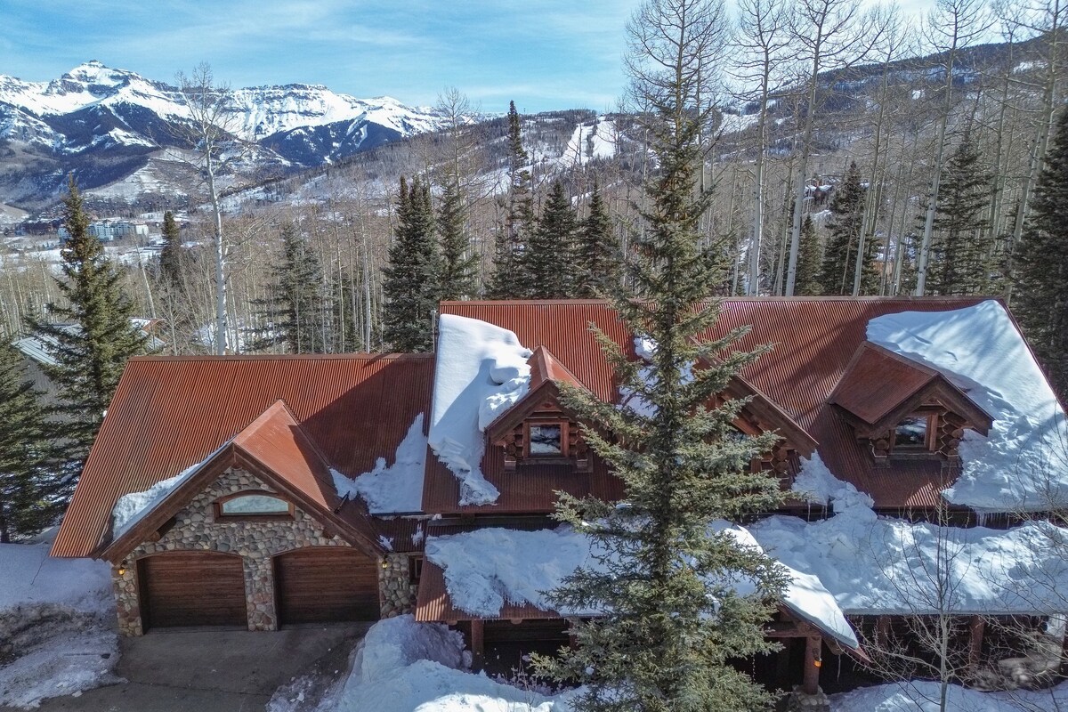 The exterior of a spacious lodge is shown, featuring a rust-colored metal roof and stone accents. Snow-covered trees surround the property, and stunning mountain views are visible in the background, indicating a serene mountain setting.