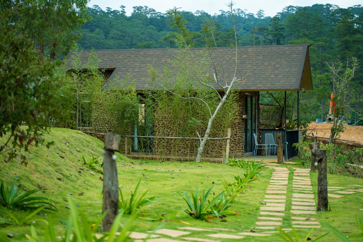 A cottage constructed with natural stone sits amidst lush greenery, surrounded by trees and rolling hills. A stone pathway leads to the entrance, bordered by well-maintained grass and shrubbery, showcasing a serene and inviting outdoor environment.