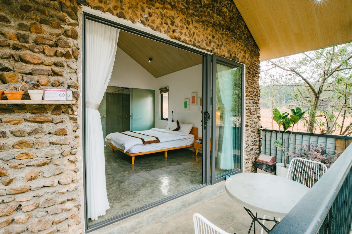 A cozy balcony area features a round table and two white chairs, providing a space to relax and enjoy the serene surroundings. Inside, a spacious bed is visible against a backdrop of natural stone walls, with soft curtains framing the large glass doors that lead to the balcony.