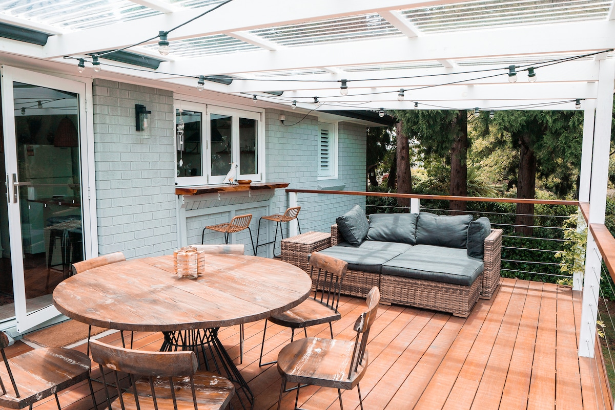 A spacious outdoor balcony is showcased with a wooden table surrounded by six metal chairs. A comfortable sofa occupies one side, complemented by cozy cushions. The area is covered with a transparent roof allowing natural light, enhancing the inviting setting among nearby greenery.