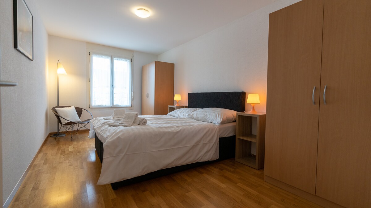A bright and spacious bedroom is presented with wooden flooring and light walls. A double bed is positioned against one wall, with bedside tables and lamps on either side. A wardrobe and a cozy chair provide additional furnishings, and natural light enters through the window.