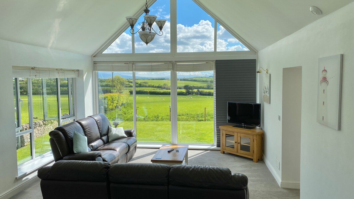A light-filled lounge area features a large sectional sofa facing broad windows with expansive rural views. A wooden coffee table sits in the center, while a television is positioned on a nearby cabinet. The high ceiling contributes to the airy atmosphere.