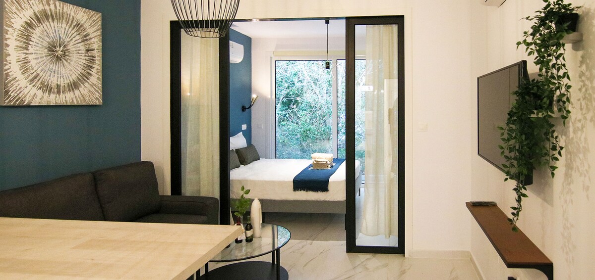 The image displays a modern living area, featuring a dark sofa and a wooden dining table beside a round glass coffee table. A spacious bedroom is visible through glass doors, showcasing a bright bed with a decorative throw. Natural greenery can be seen outside the windows.
