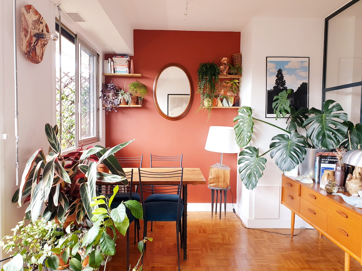 A dining area features a wooden table accompanied by four black chairs. On the wall, a round mirror is hung above floating shelves filled with plants and decorative items. A floor lamp stands next to a cabinet, which is adorned with various plants and art pieces.