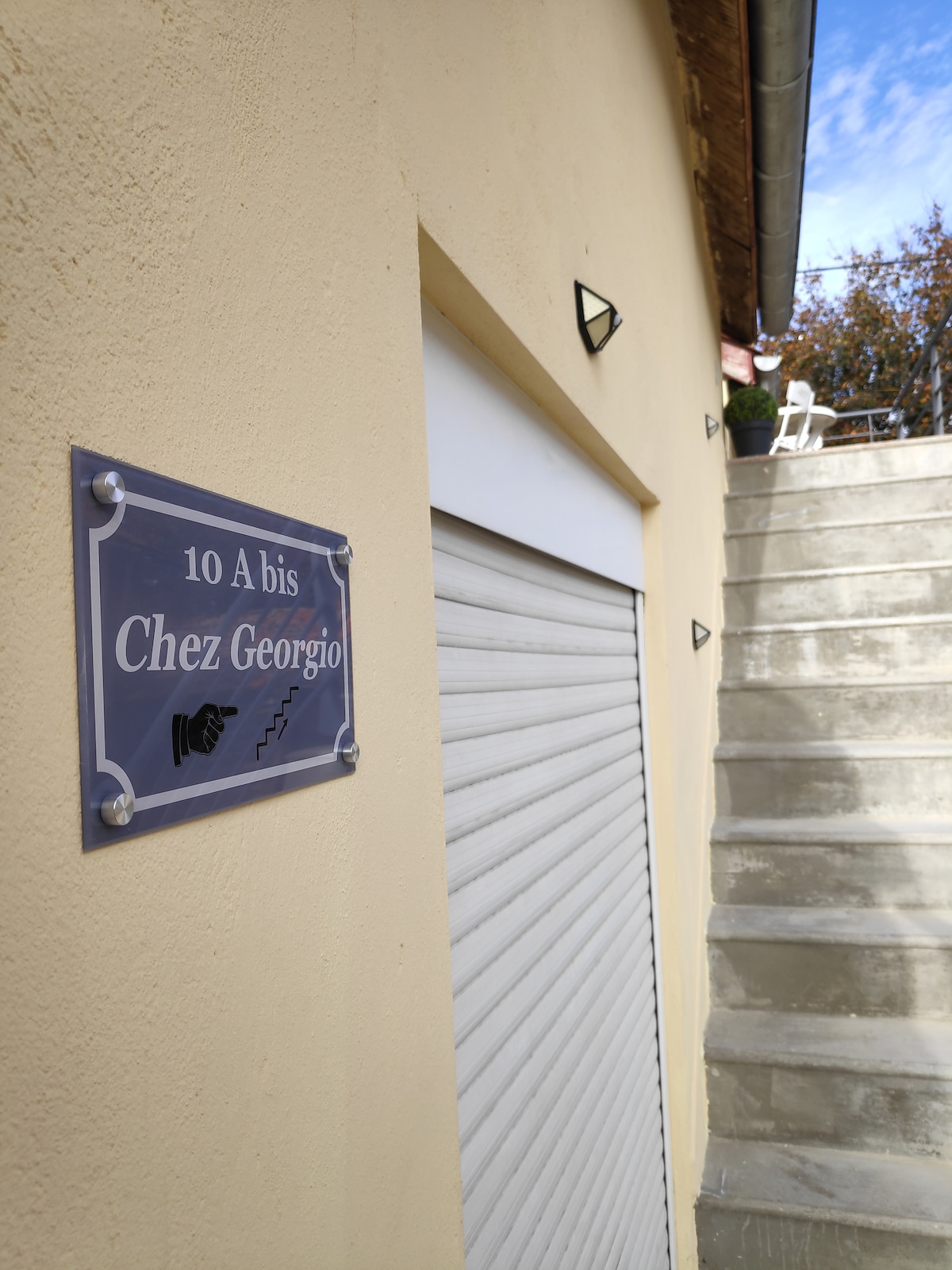 The image features a beige exterior wall, showcasing a sign that reads '10 A bis Chez Georgio.' A set of concrete stairs leads upward, accompanied by a closed garage-style door. Subtle architectural details, such as metal fixtures, are also visible.