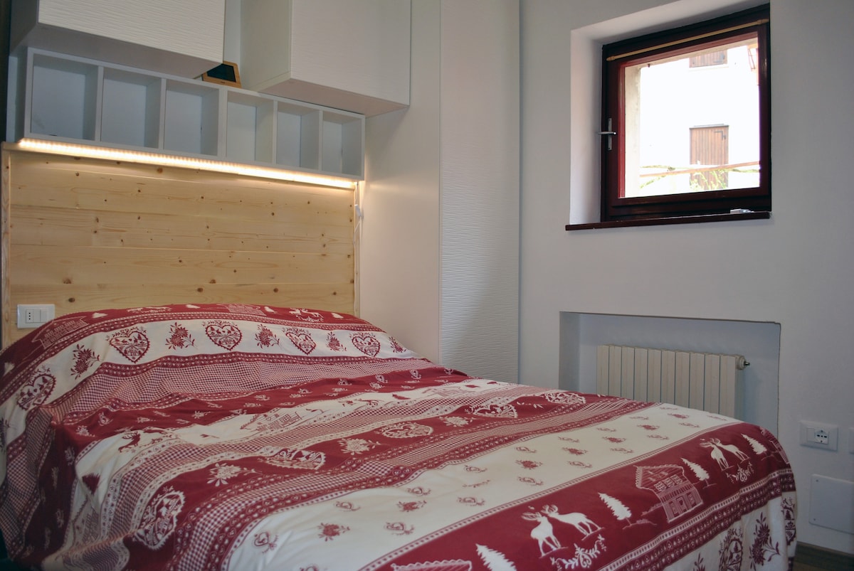 A cozy sleeping area features a comfortable bed draped in a patterned red and white bedspread. A wooden headboard with soft lighting adds a warm touch. A small window allows natural light to enter, complemented by a radiator beneath.