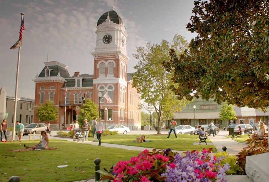 A historic red-brick building is featured, showcasing its tall clock tower. A green park area in the foreground includes lush grass, blooming flowers, and people engaging in various outdoor activities. Trees provide shade and a communal atmosphere under a soft blue sky.