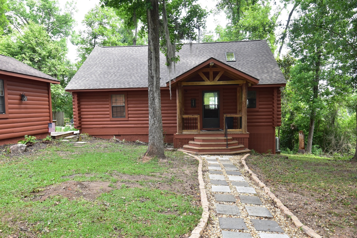 The exterior of a rustic log cabin is displayed, featuring a brown wooden facade and a welcoming front porch. A stone pathway leads to the entrance, bordered by well-kept grass and trees. Natural surroundings create a serene atmosphere.
