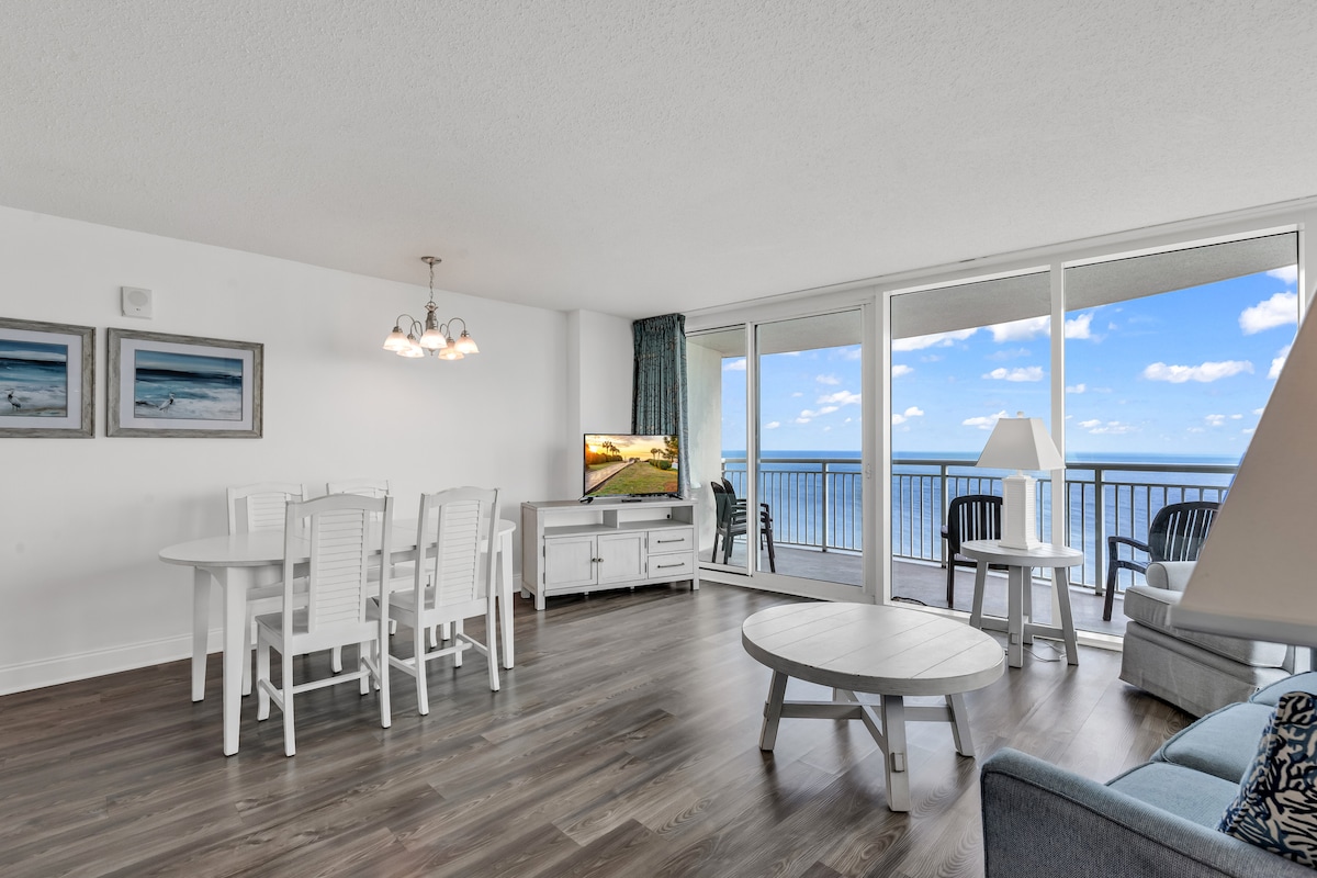 The living area features large glass doors that offer extensive ocean views, allowing natural light to fill the space. A round coffee table and white dining set are present, complemented by a cozy seating area. A flat-screen TV is positioned for easy viewing.