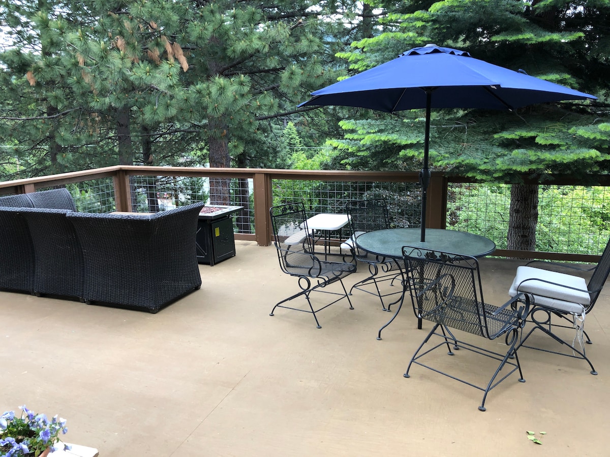 An outdoor deck features a seating area with wicker furniture complemented by a blue umbrella. A circular table with metal chairs is positioned next to the seating, surrounded by lush greenery from tall trees, creating a peaceful outdoor space.