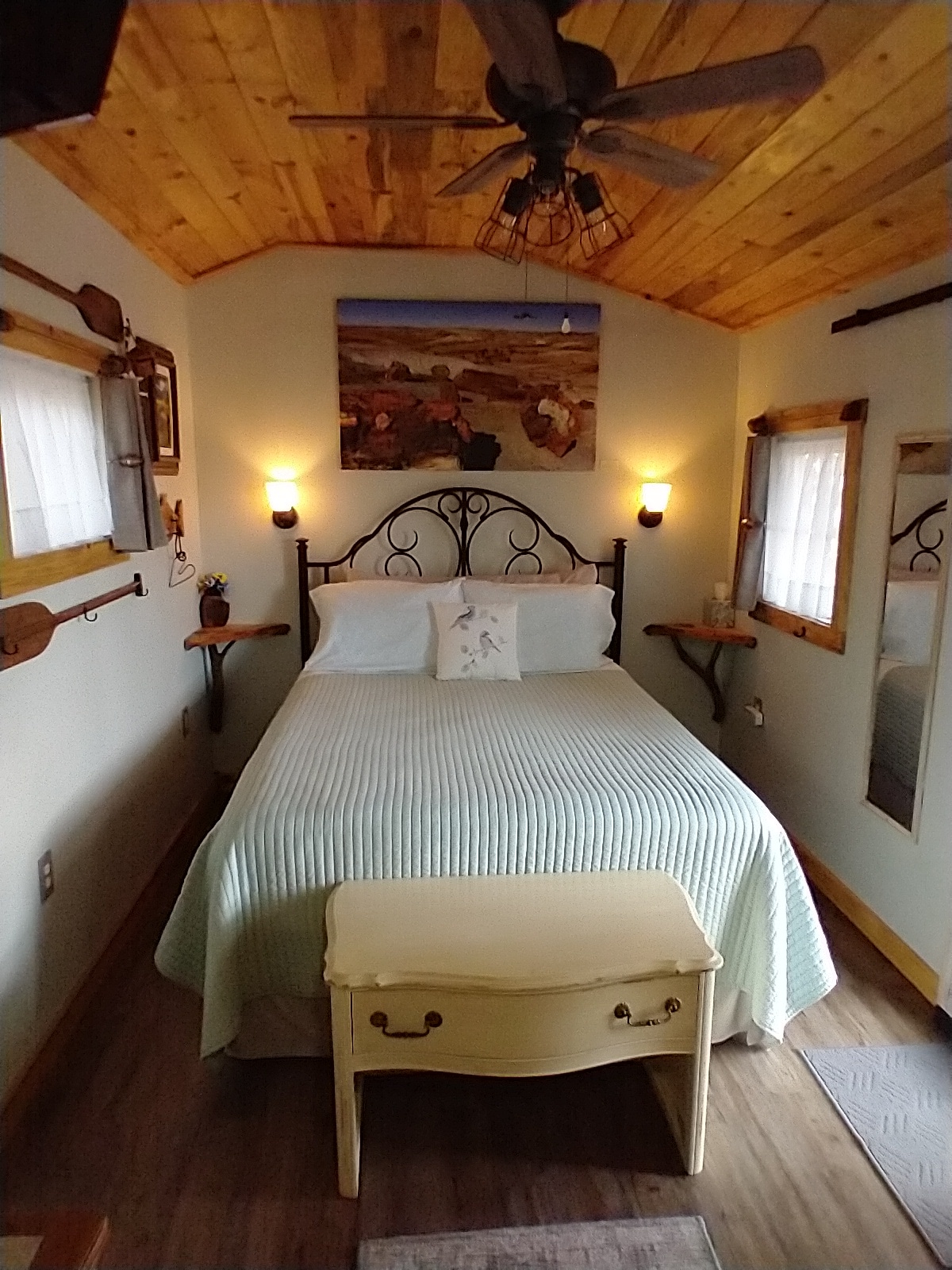 A cozy sleeping area features a comfortable bed with a light green quilt and decorative pillows. Wooden walls and ceiling create a warm ambiance, while wall sconces and natural light from windows enhance the space. A small side table and a mirror contribute to the room's rustic charm.