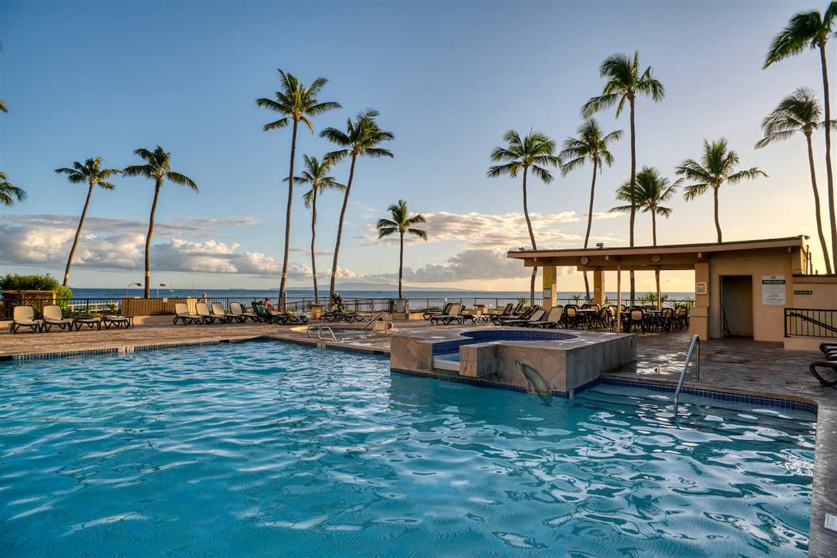 A sparkling pool is framed by palm trees, with a hot tub positioned nearby. Lounge chairs are arranged around the pool area, and a gazebo is visible in the background, providing shaded seating. The tranquil ocean can be seen beyond the pool.