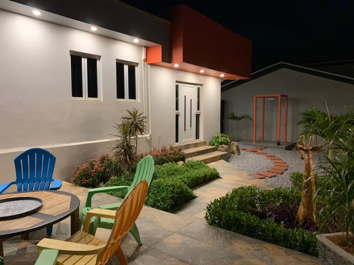 An inviting outdoor area is showcased, featuring a stone pathway leading to the entrance. Colorful chairs surround a circular table, complemented by landscaped greenery and vibrant flowering plants. Soft lighting illuminates the facade of the villa, enhancing the welcoming atmosphere.