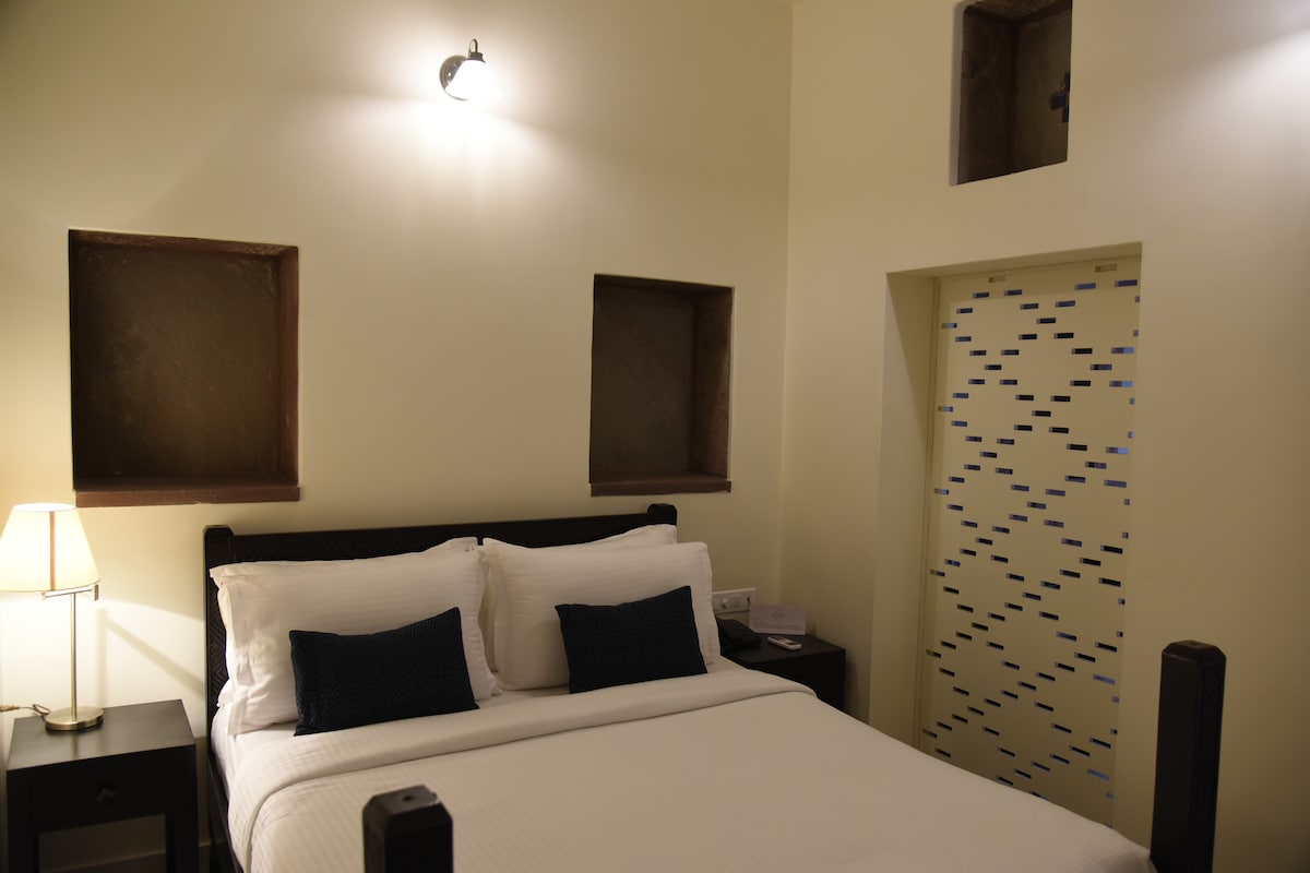 A well-lit sleeping area features a spacious bed with white bedding and two dark cushions. Simple nightstands are placed on either side, and the walls are adorned with small windows, providing a glimpse of the building's heritage. A patterned feature wall adds interest.