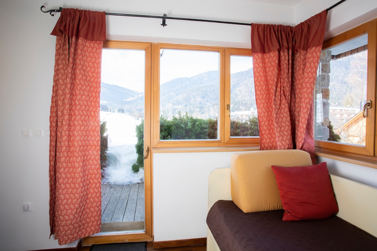 A cozy corner of the living space is highlighted by large windows framed with rich, patterned curtains. The view showcases the serene outdoors, with mountains visible in the distance. A simple sofa with a colorful cushion complements the inviting atmosphere.