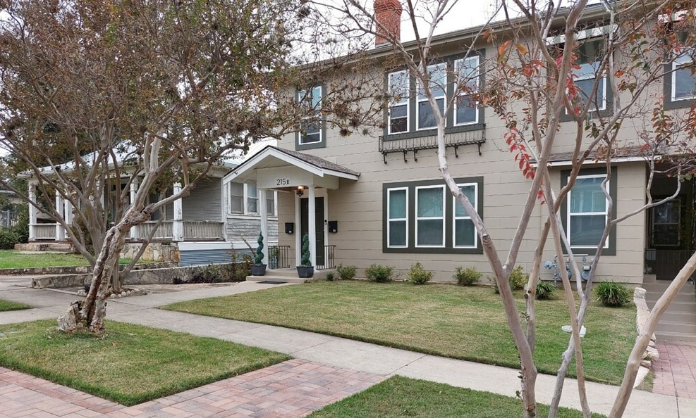The exterior of a duplex building is presented, featuring a welcoming entrance with a front porch. A well-maintained grassy area is visible in front of the property, alongside paved walkways. Large windows provide natural light and a sense of openness.