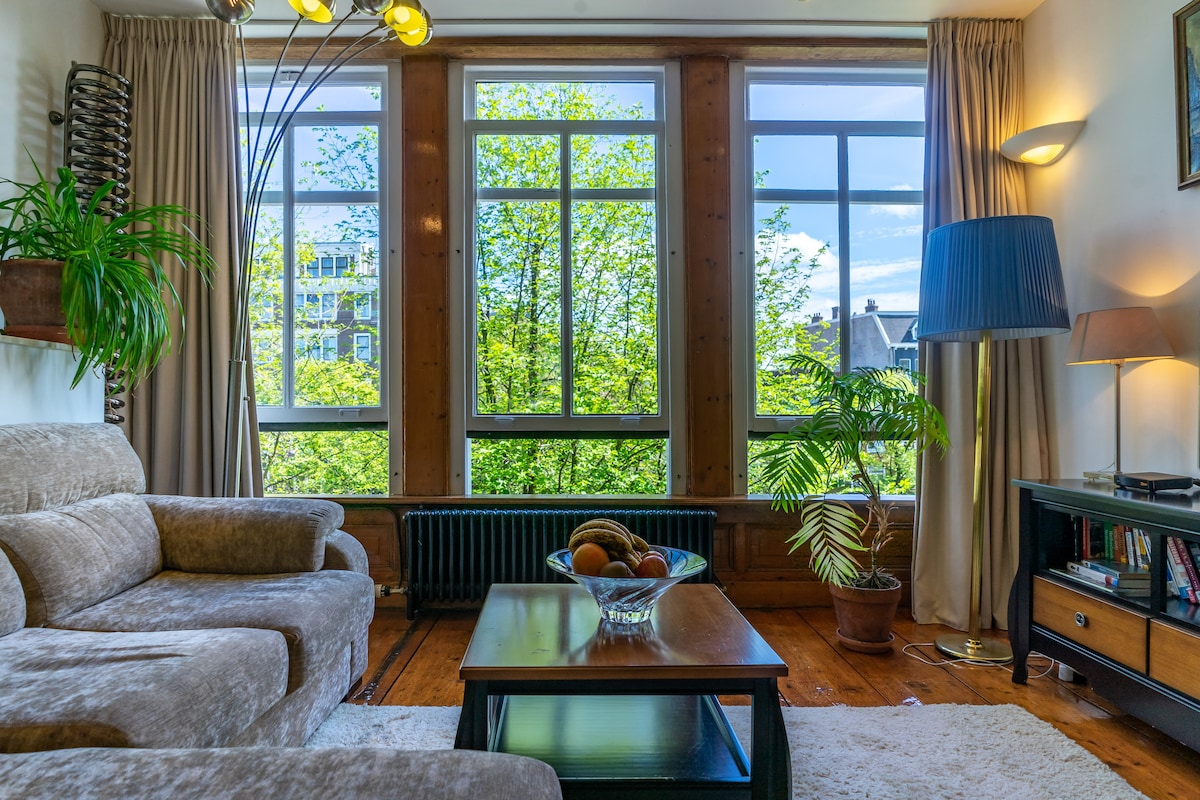 Natural light fills the spacious lounge area through large windows with a view of greenery outside. A comfortable sofa complements the wooden accents. A coffee table with a bowl of fruit is centered on a soft rug, while potted plants add a touch of freshness.