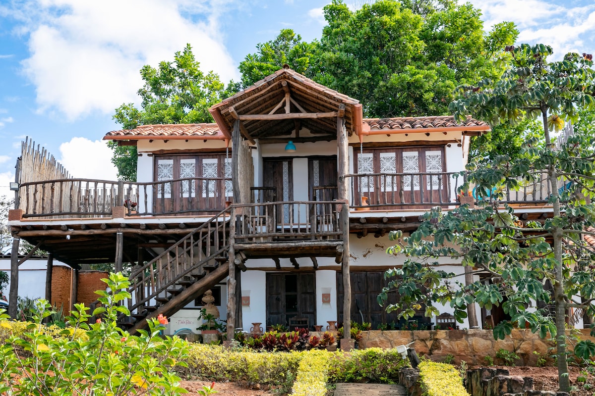 A charming two-story structure features a wooden balcony with decorative railings, surrounded by lush greenery. Multiple windows allow natural light into the building. Steps lead up to the entrance, framed by intricate wooden details and vibrant plants lining the pathway.