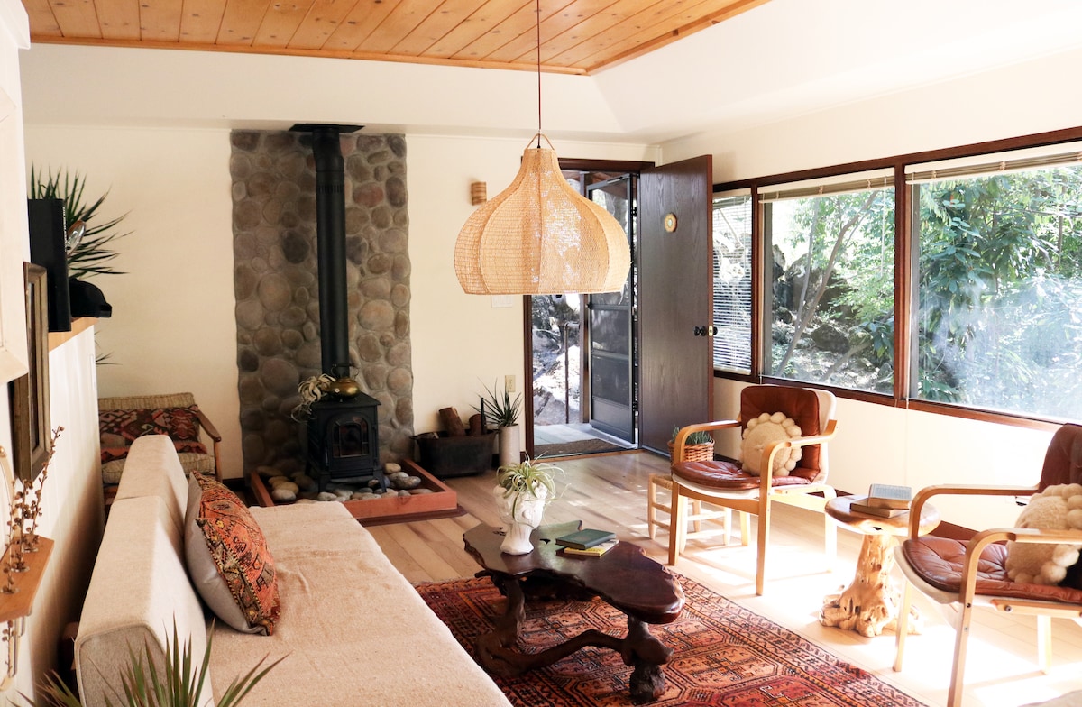 A bright and inviting living area features large windows that frame lush greenery. A stone fireplace adds a cozy focal point, while carefully arranged seating creates a comfortable space. Natural wood elements and textile accents contribute to the cabin's charming ambiance.