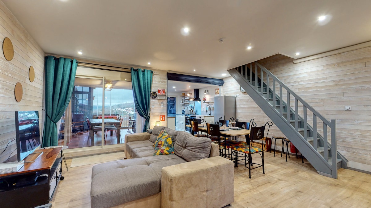 A spacious living area features warm wooden accents and large windows that provide ample natural light. A comfortable sectional sofa and a dining table with several chairs are positioned within the space. A staircase leads to the upper level, adding dimension to the open layout.