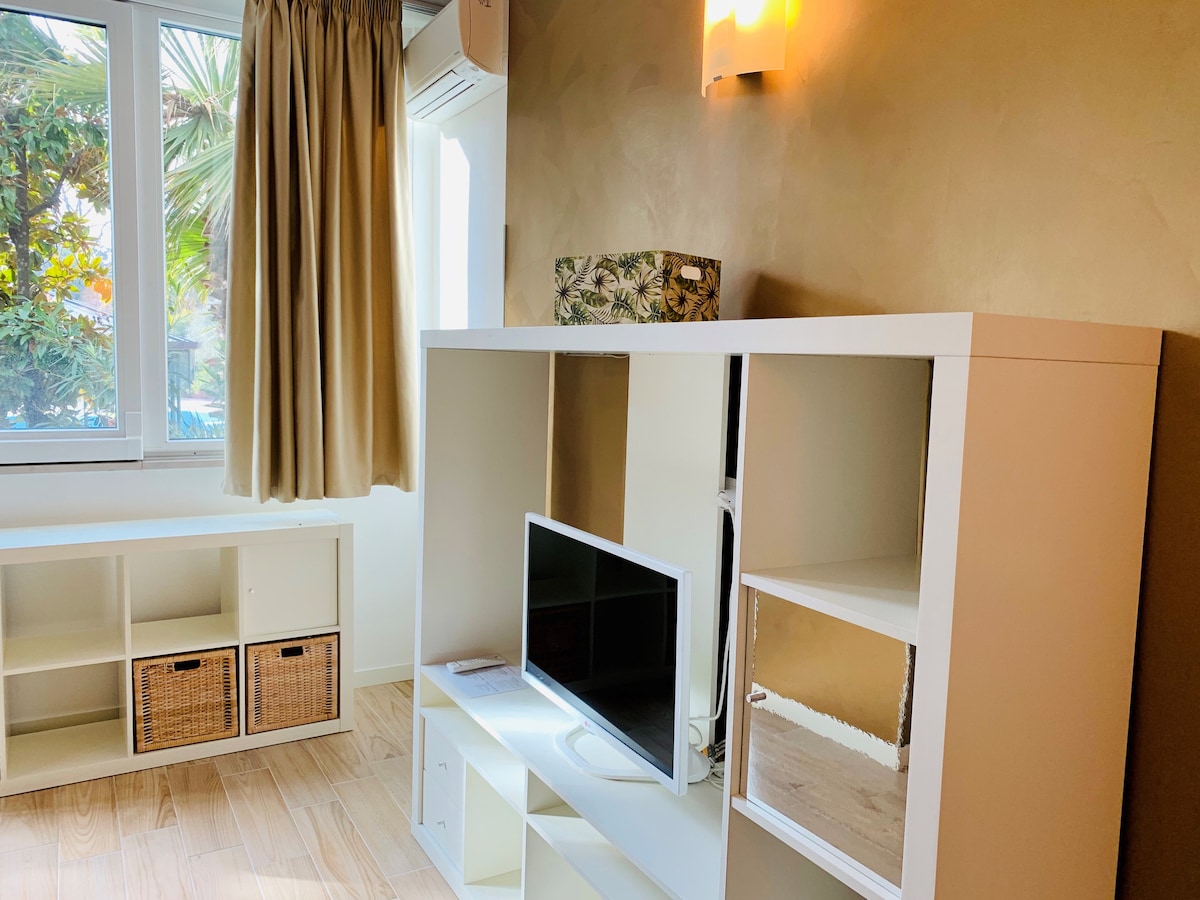 A light-filled living area features a white entertainment unit with a television, complemented by a storage shelf containing woven baskets. Large windows with sheer curtains allow natural light to illuminate the space, giving a sense of openness.