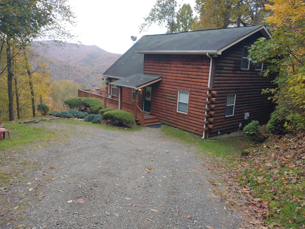 A log cabin sits nestled against a backdrop of autumn foliage, showcasing warm wood tones. The sloping gravel driveway leads to the entrance, while a spacious porch extends along the front, offering scenic views of the mountains in the distance.