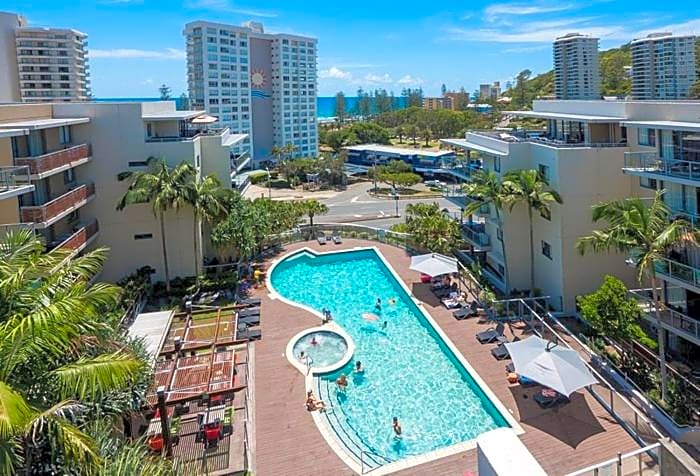 The image showcases a spacious heated pool surrounded by palm trees and lounge chairs. A hot tub is visible by the poolside. In the background, a view of the ocean and nearby buildings adds to the scene, under a clear blue sky.