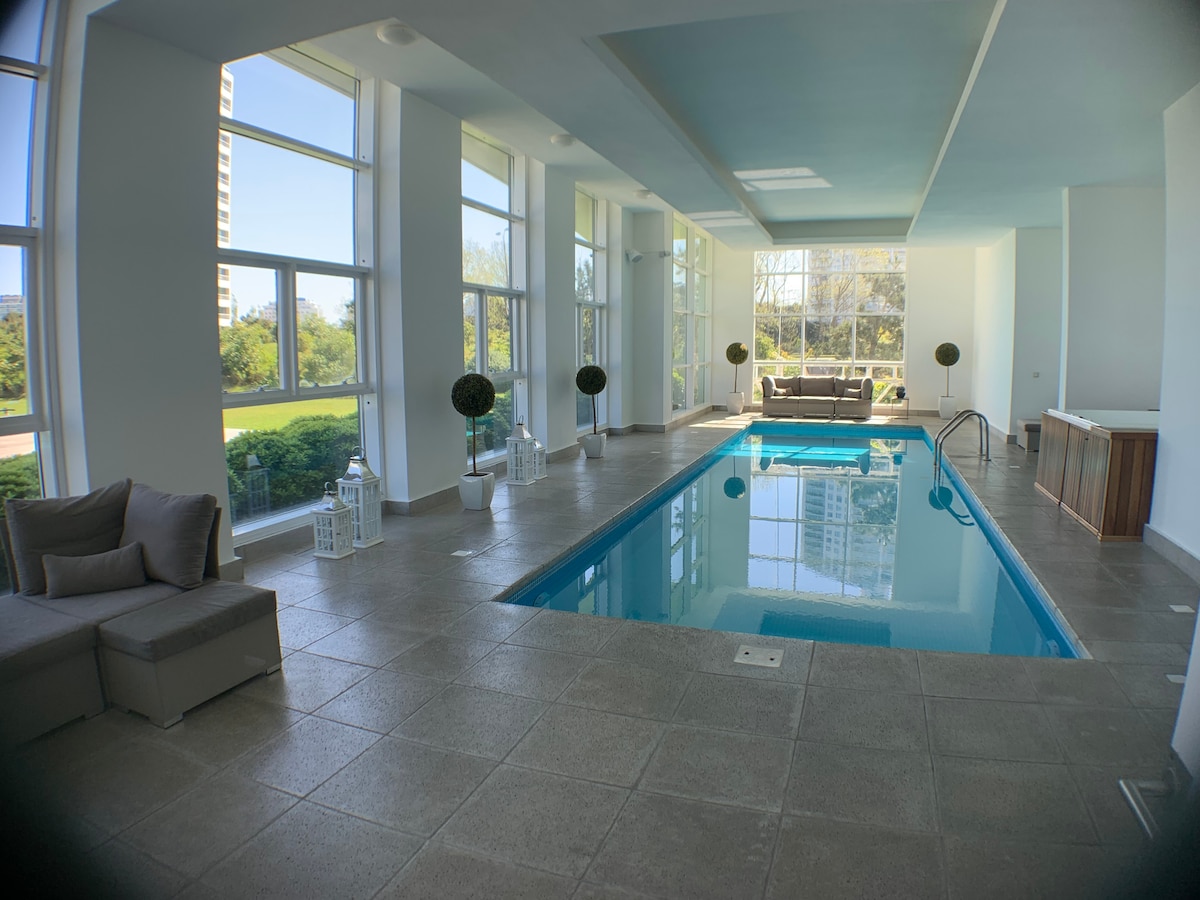 An indoor pool area is illuminated by large windows, offering views of the surrounding greenery. Comfortable seating is arranged nearby, while elegant lanterns add to the calm ambiance. The space is characterized by a modern design, with a tranquil blue ceiling accentuating the serene environment.