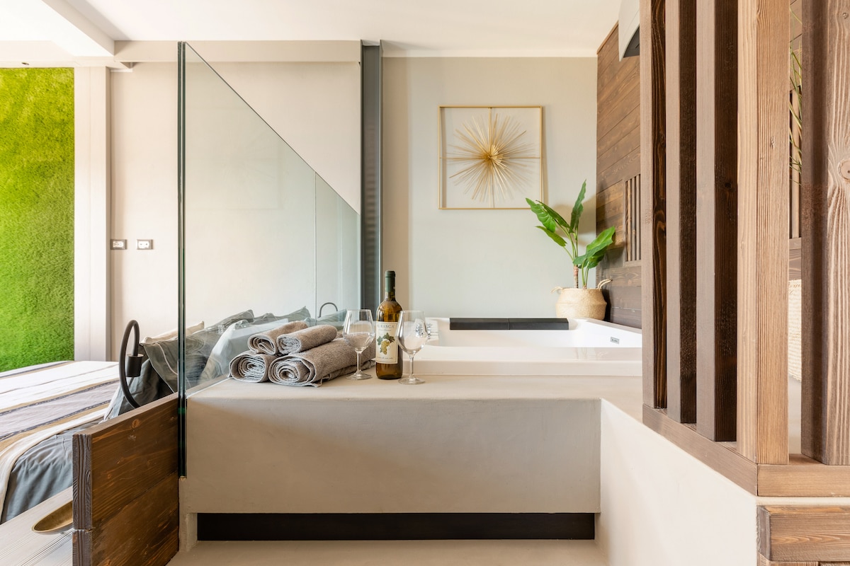 A modern bathroom is showcased, featuring a spacious jacuzzi and a sleek glass partition. Soft, rolled towels are neatly arranged next to a bottle of wine, set on a muted countertop. A green plant adds a touch of nature to the contemporary design.