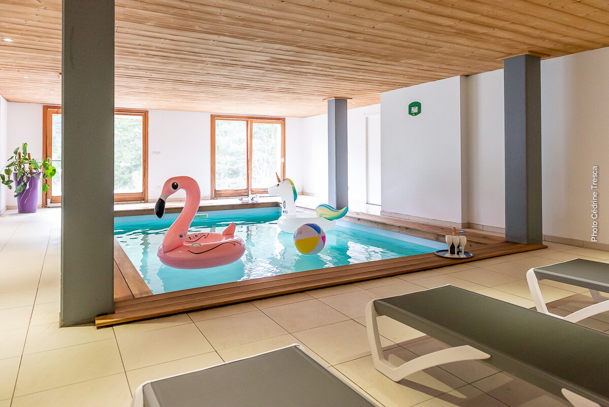 An indoor heated swimming pool features colorful inflatable toys floating on the water. Sunlight filters in through large windows, illuminating the space. Lounge chairs are positioned nearby, creating a relaxed atmosphere for leisure activities.
