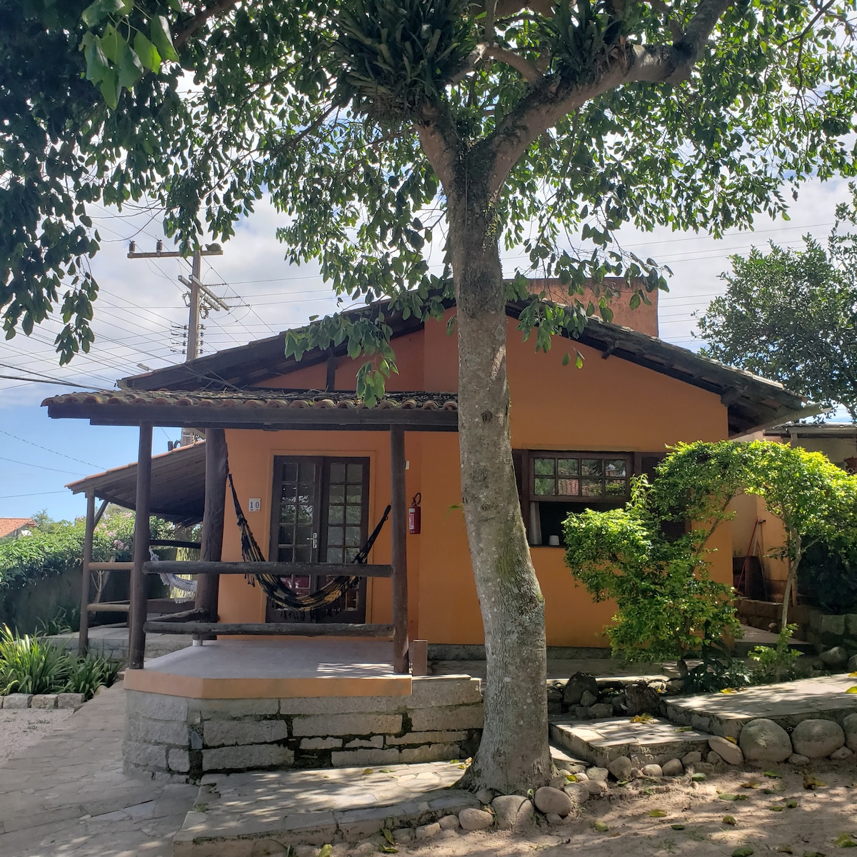 The exterior of a charming cabin is shown, featuring warm orange walls and a welcoming porch. A hammock hangs nearby, and lush greenery surrounds the entrance. Natural stone pathways lead to the cabin, enhancing its inviting presence in a tranquil outdoor setting.