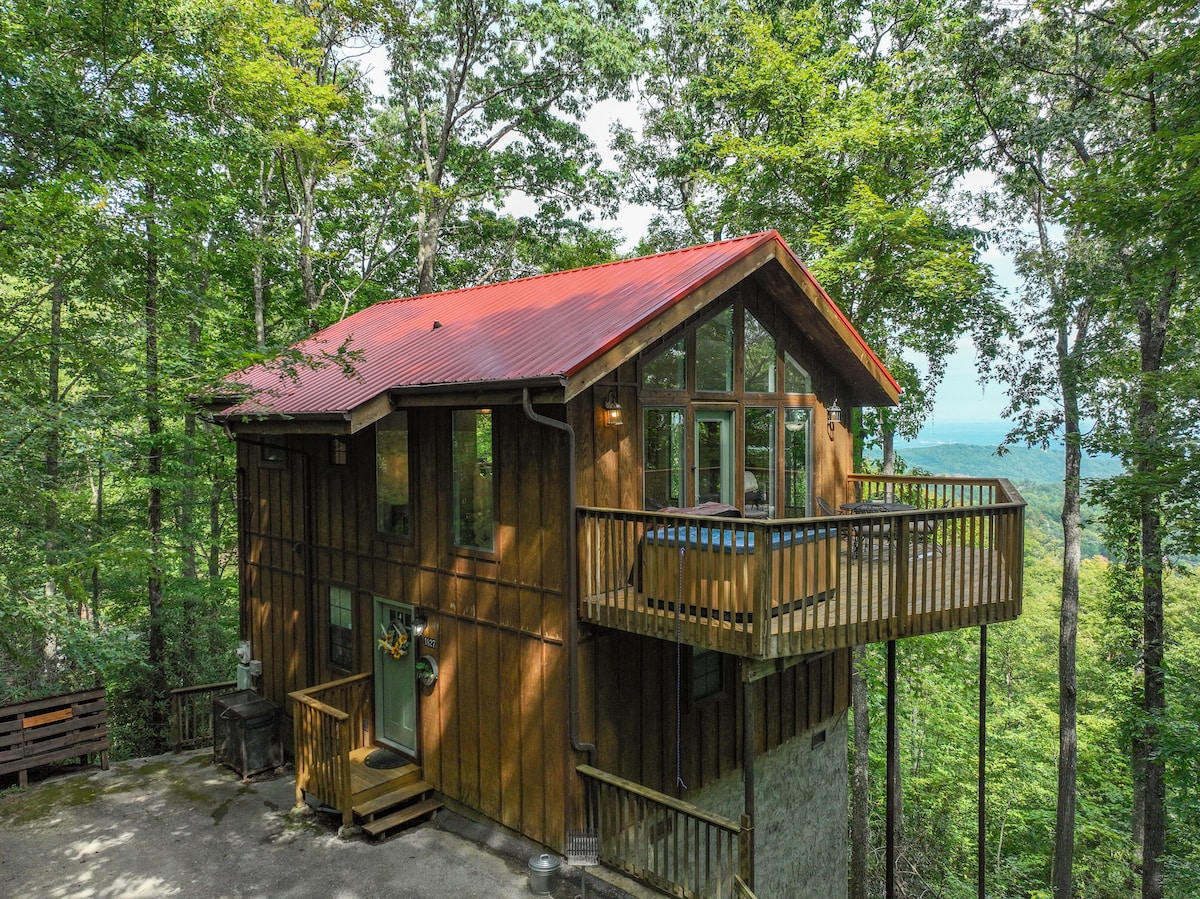 A cabin nestled among trees features a red metal roof and a wooden exterior. Large windows allow natural light to fill the interior, while a private balcony extends from the upper level, offering serene views of the surrounding forest and mountains.