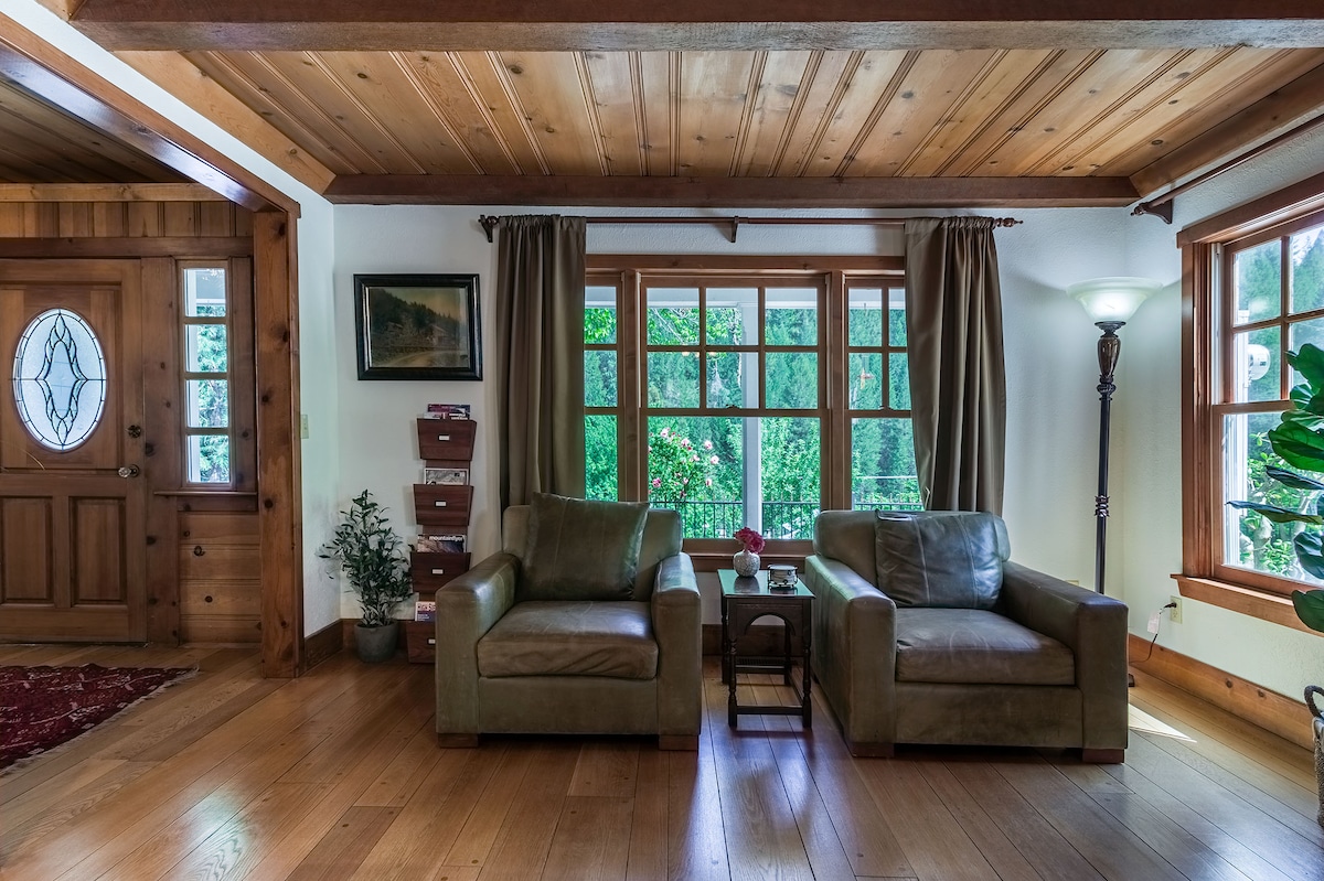A cozy living room features two leather armchairs positioned beside a small coffee table. Large windows are adorned with dark curtains, allowing natural light to illuminate the wood flooring. A potted plant and a welcoming entryway are visible, contributing to a warm environment.