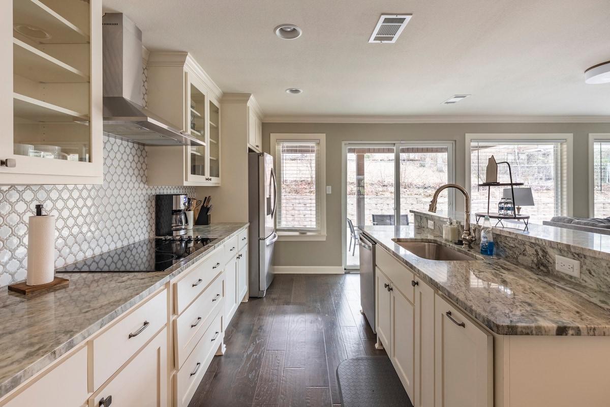 A spacious kitchen showcases light-colored cabinetry and a large island with a countertop made of granite. Stainless steel appliances are present, alongside ample natural light coming from multiple windows. The open layout connects to a living area, enhancing the overall functionality of the space.