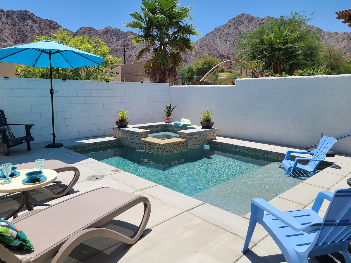 A private dipping pool features a Cabo Shelf for lounging and a spillover waterfall from the adjacent hot tub. Surrounded by a privacy wall, the area includes lounge chairs, a small table set for outdoor dining, and palms framing the stunning mountain views.
