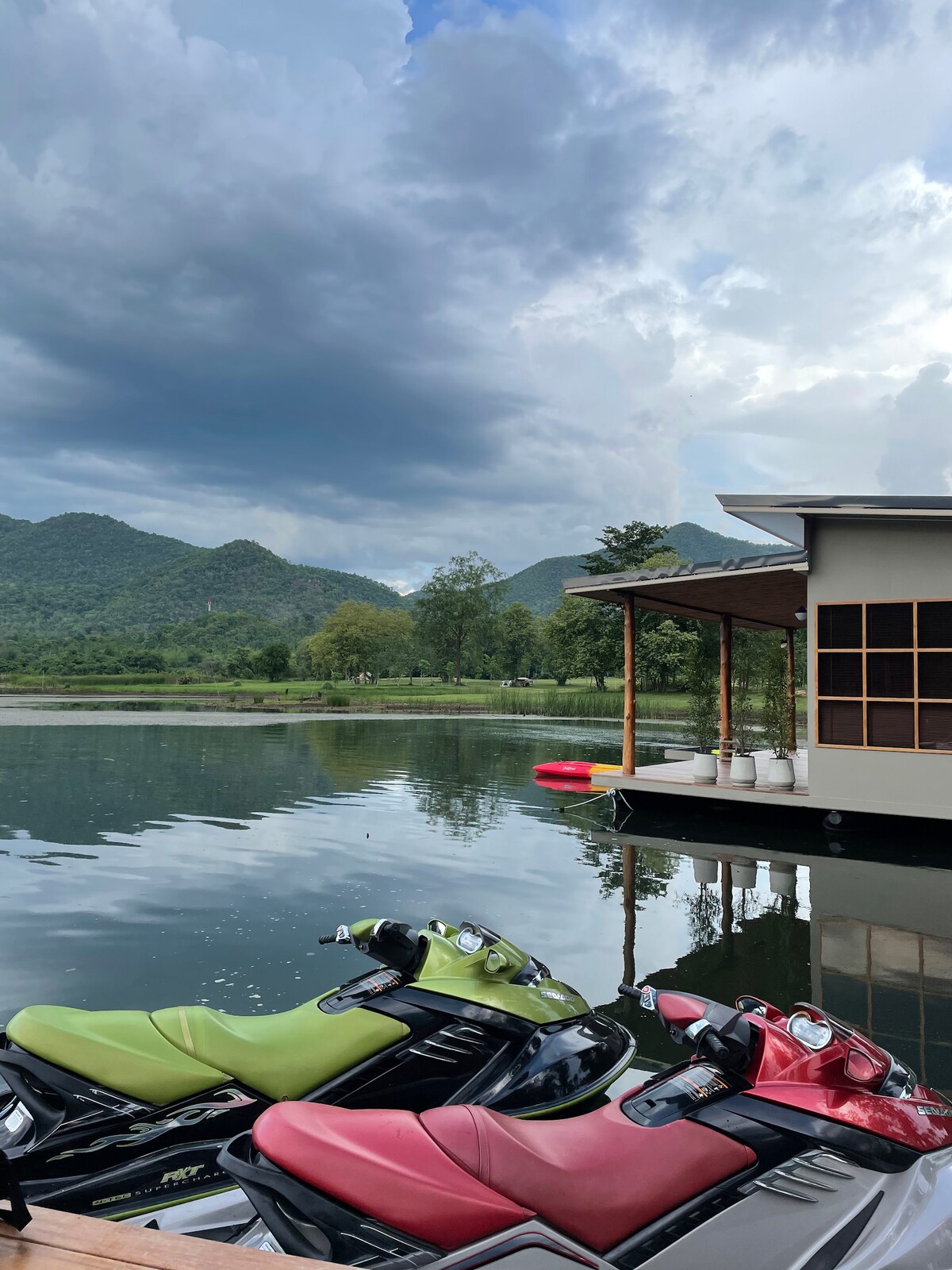 A serene lake view features floating villas nestled against the backdrop of green mountains. Reflections shimmer on the water's surface, while a red inflatable and several jet skis, in vibrant colors, are docked along the edge, indicating opportunities for leisure activities.