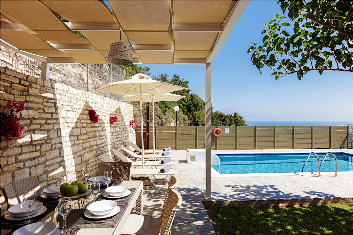An outdoor dining area is presented under a shaded pergola, featuring a dining table set for multiple guests. The background showcases a private swimming pool surrounded by sunbeds, umbrellas, and lush greenery. The clear blue sky extends above, highlighting the tranquil atmosphere.