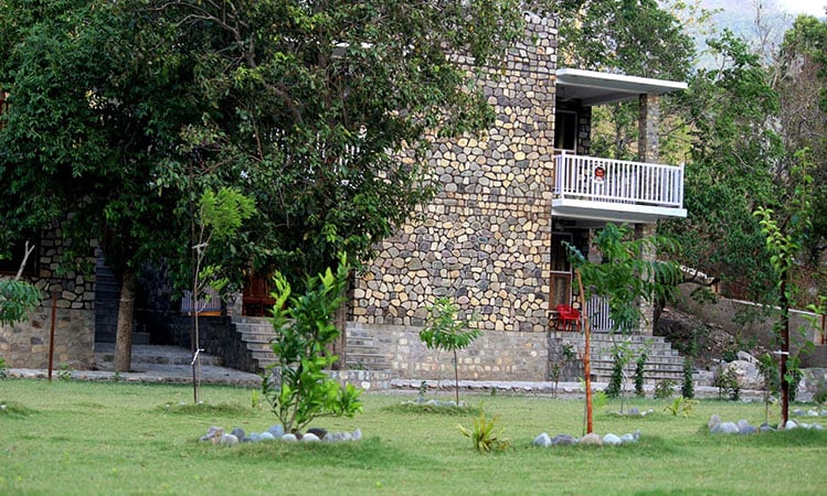 The exterior of the resort features a stone structure surrounded by lush greenery. A balcony overlooks the spacious lawn, which includes several small trees and decorative stone arrangements. The natural landscape creates a serene environment, ideal for relaxation.