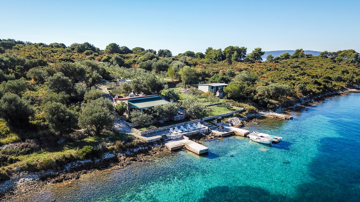 A scenic aerial view captures the serene seaside landscape, featuring green trees surrounding two structures. A private boat is anchored near a stone pier, while sunbeds are arranged along the shoreline. Clear blue waters gently lap against the rocky coast.
