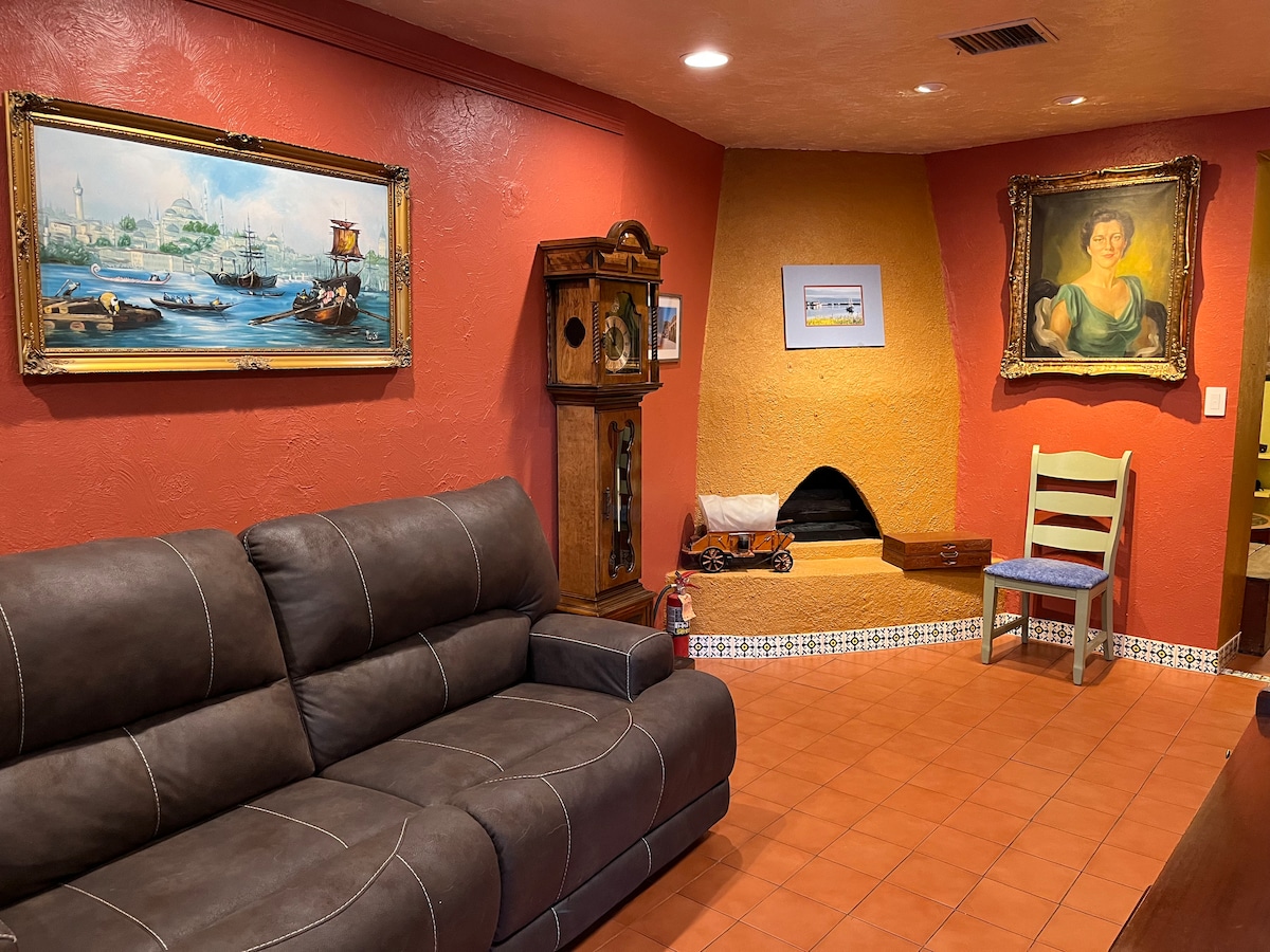 A cozy sitting area features a brown sectional sofa and a colorful wall adorned with framed artwork. A decorative wooden clock and a unique fireplace create an inviting focal point, while terracotta floor tiles add warmth to the space.