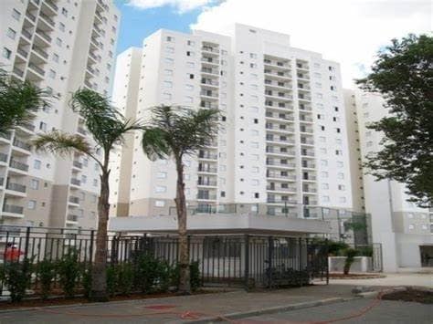 A modern condominium is depicted with multiple high-rise buildings surrounded by landscaped greenery. Tall palm trees line the entrance, which is secured by a wrought-iron gate. The skyline showcases a clear blue sky, contributing to an open and inviting atmosphere.