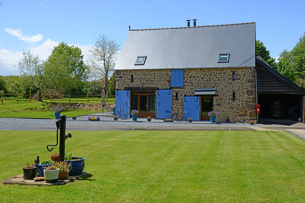 A charming barn with stone exterior and blue window shutters sits on a well-manicured lawn. A vintage water pump and flower pots add character in the foreground, while trees and a farm area are visible in the background under a clear blue sky.