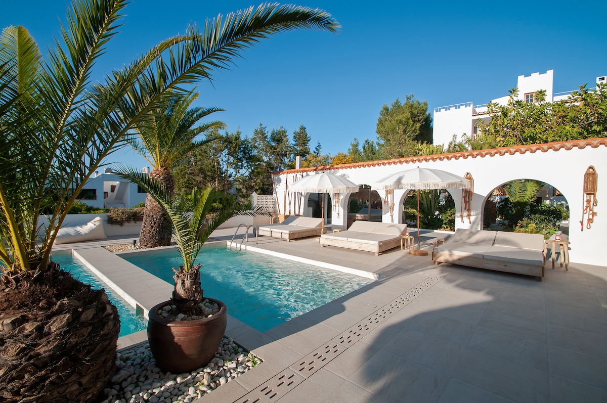 An inviting pool area is surrounded by palm trees, with two sun loungers positioned near the shimmering water. A shaded seating area with white canopies provides comfort, and neat landscaping enhances the overall serene environment. The clear blue sky complements the tranquil setting.