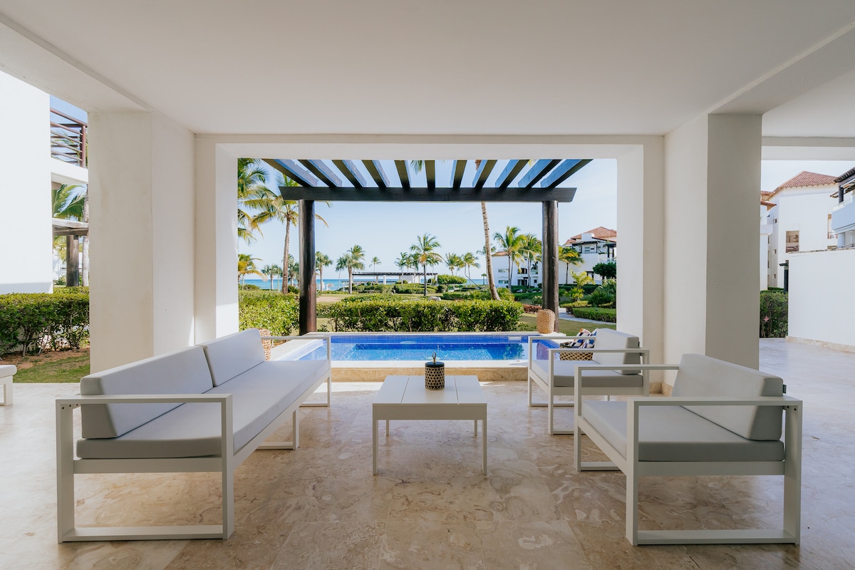 A shaded terrace area is presented with modern white seating and a small coffee table, overlooking a private mini pool. Lush tropical gardens and the ocean are visible in the background, creating a serene outdoor space for relaxation and socializing.